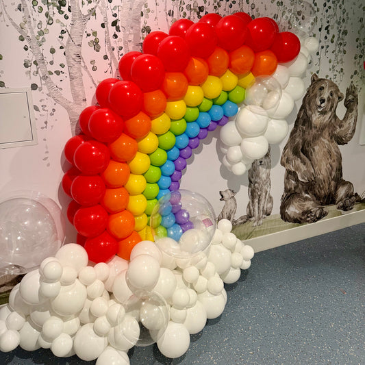 Balloon arch shaped like a rainbow with white clouds in a room setting.