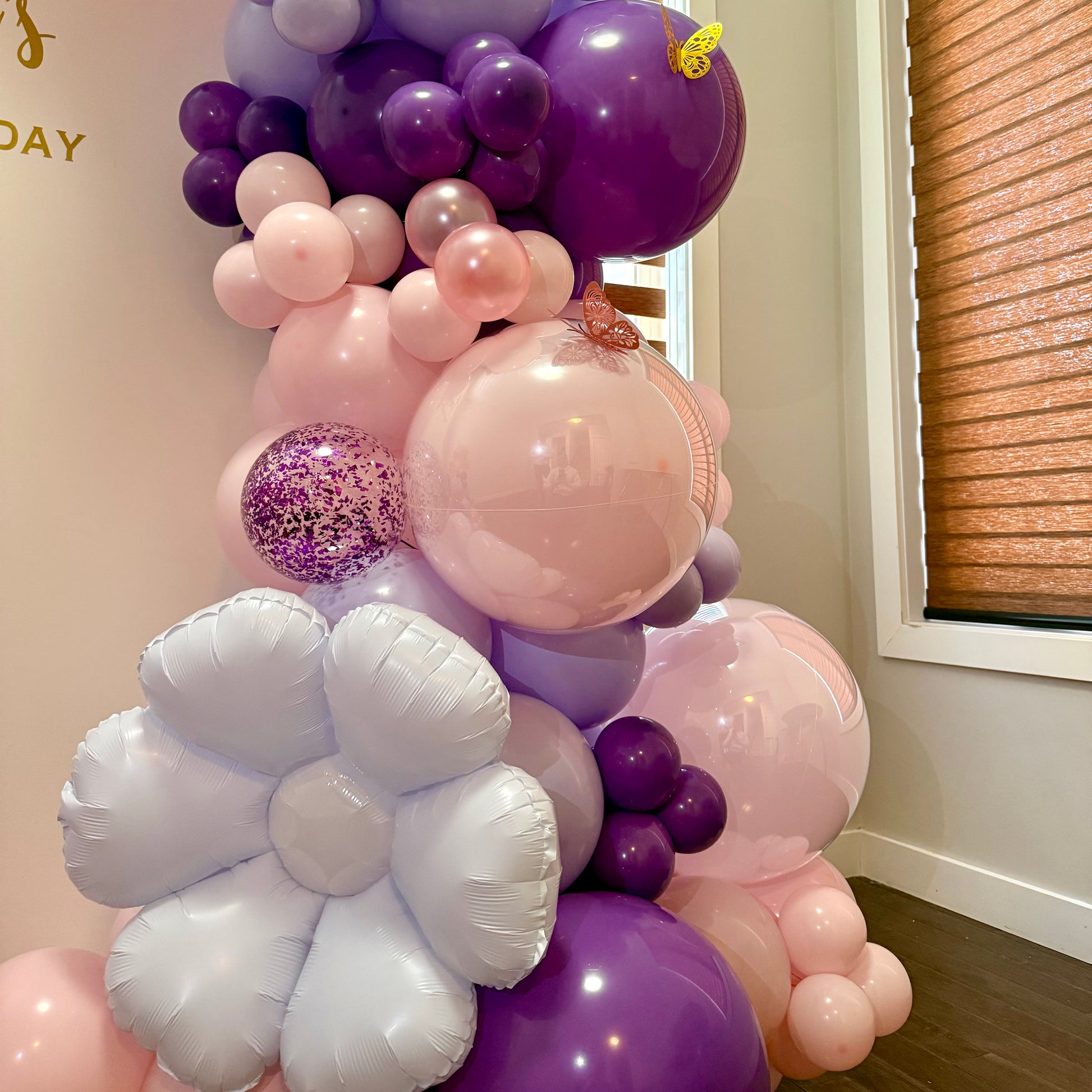 Colorful balloon arch with purple, macaroon pink, and flowers balloons against a neutral wall.