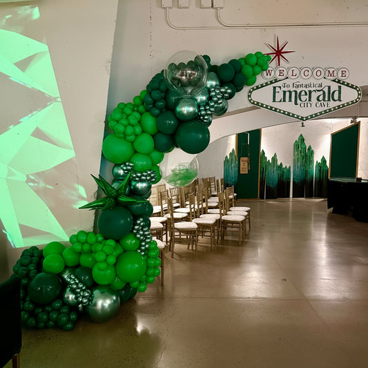 Green balloon archway with chairs and 'Welcome to the Fantastical Emerald City Cave' sign.